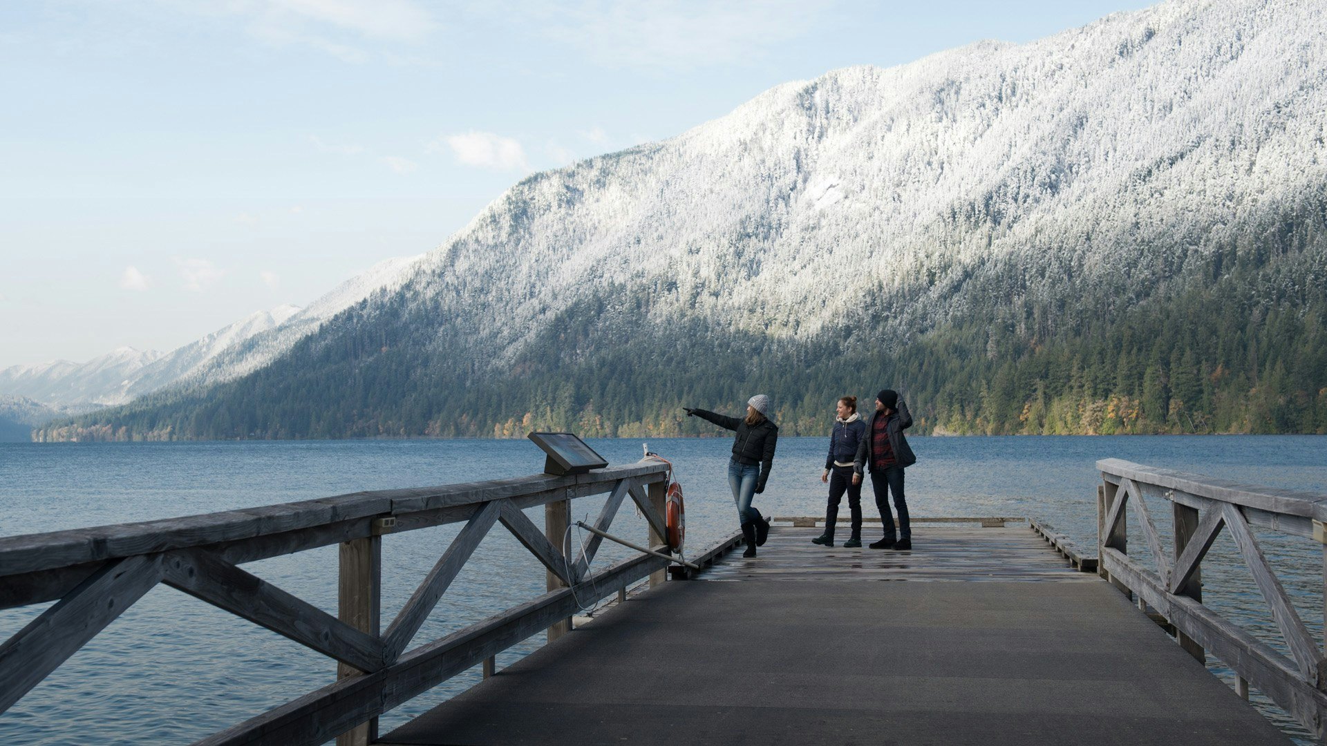 three people at the edge of a dock in front of lake and winter mountains